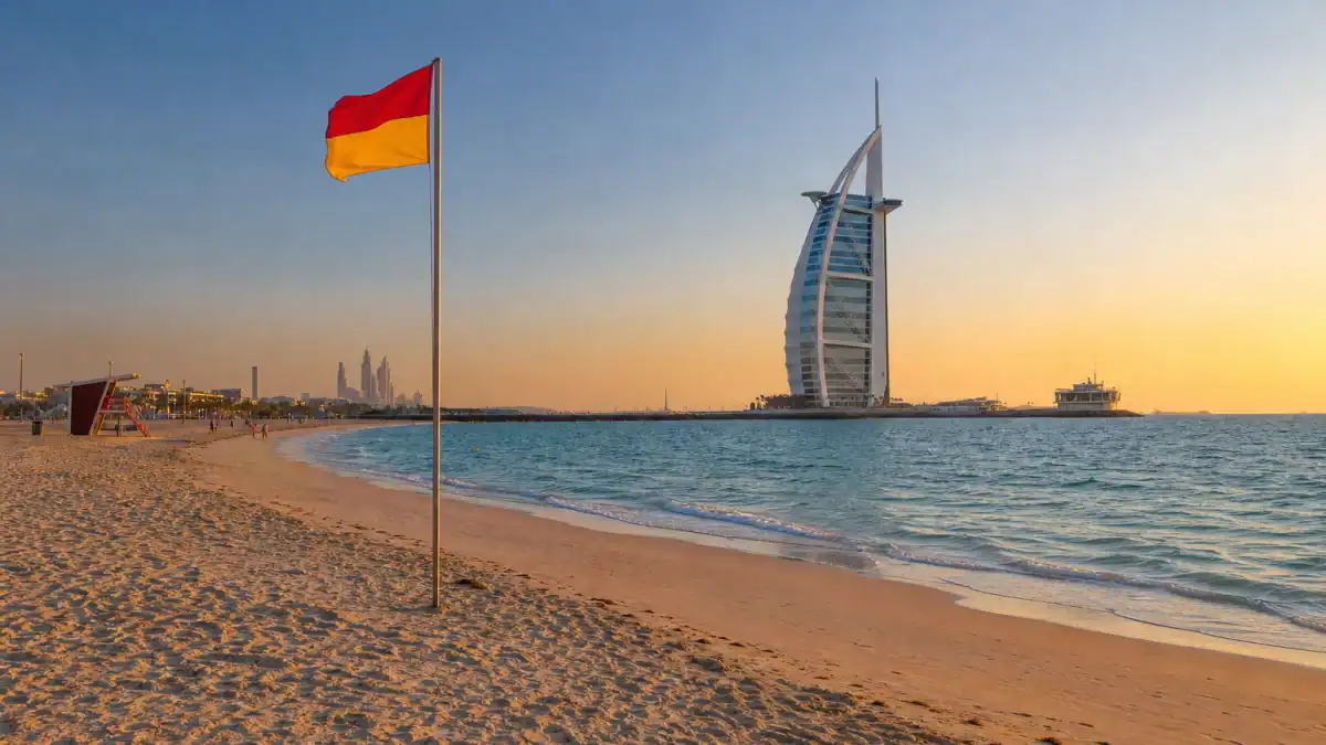 شاطئ الغروب في دبي مع برج العرب وراية السلامة قرب البحر.